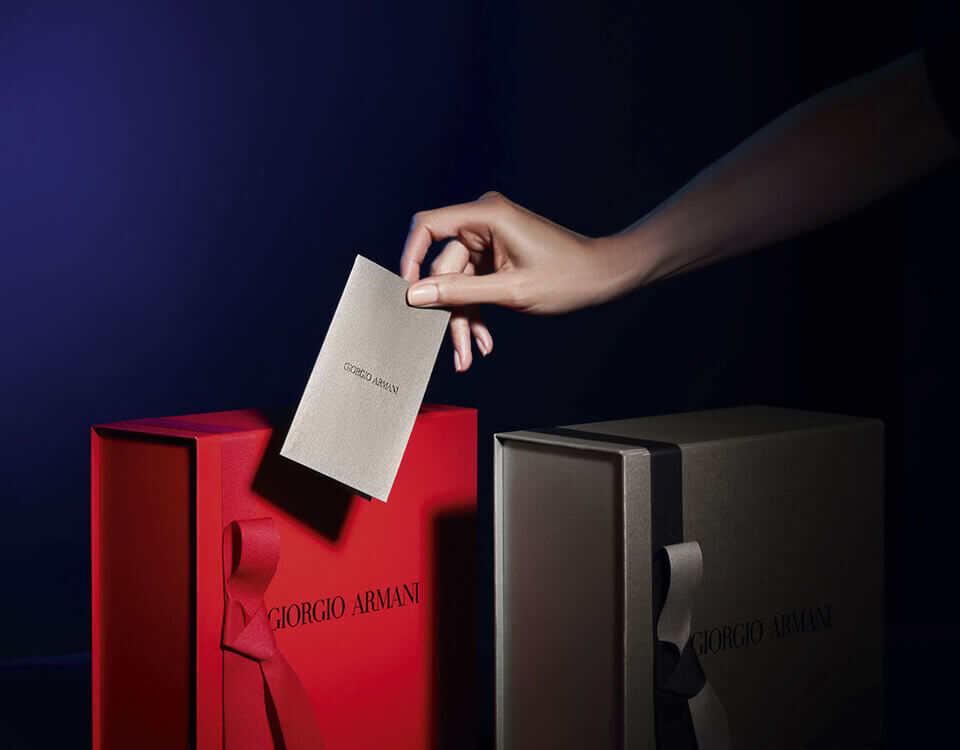 A hand places a card into a red gift box labeled "Giorgio Armani," with a green gift box beside it, both featuring elegant ribbons in a dimly lit setting.