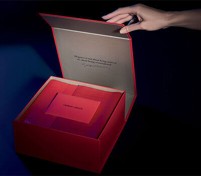 A hand opens a red gift box, revealing a neatly arranged interior with a card inside, all set against a dark background.