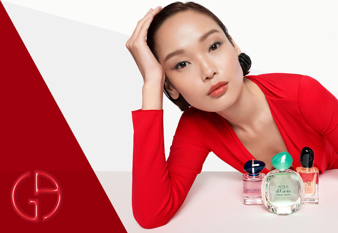 A model in a red outfit leans on her hand with a neutral expression, surrounded by various perfume bottles on a white background.