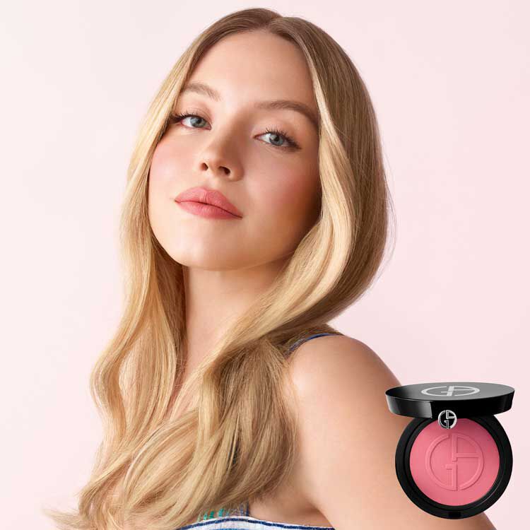 A model with long, wavy hair and a soft makeup look poses beside a round compact of blush with a sleek black case. The background features a pink gradient with blush powder scattered in a vibrant shade of pink.