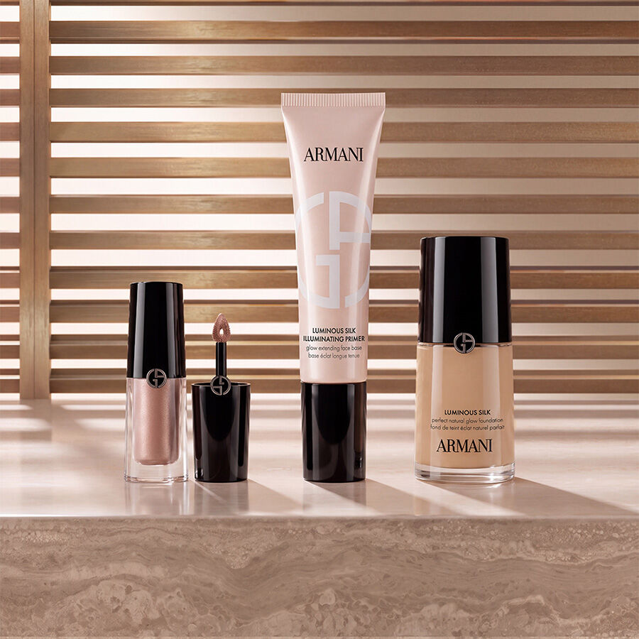 Armani beauty Make-up-Produkte, darunter Liquid Highlighter, Luminous Silk Illuminating Primer und Luminous Silk Foundation, prasentiert auf einer Marmoroberflache vor einem warmen Lamellenhintergrund.