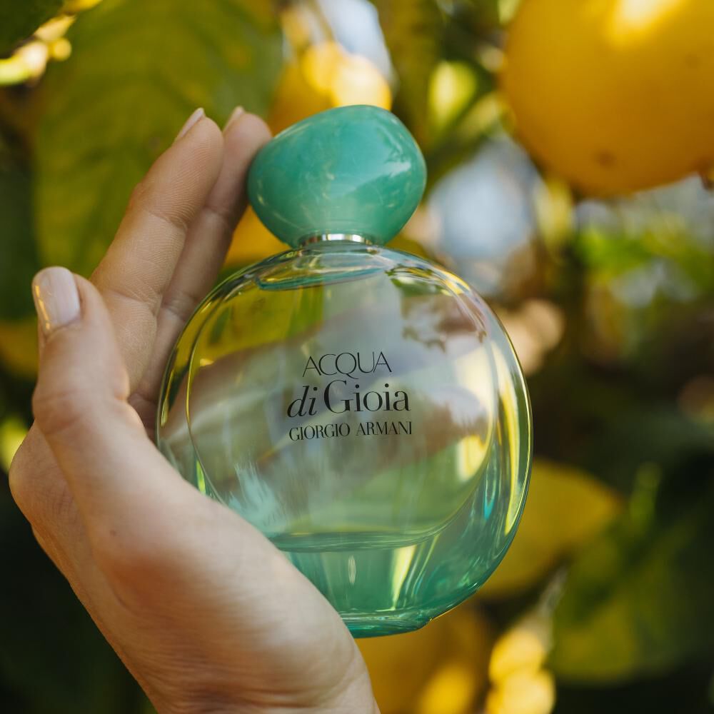A hand holding a bottle of Acqua di Gioia perfume by Giorgio Armani, set against a backdrop of vibrant green leaves and lemon fruits.
