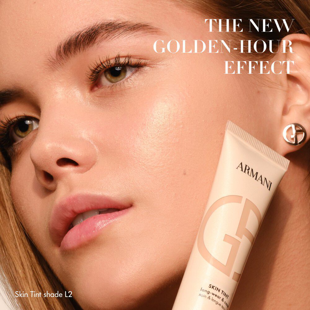 Frau mit strahlender Haut, die eine Armani Skin Tint Tube halt, mit dem Text THE NEW GOLDEN-HOUR EFFECT.
