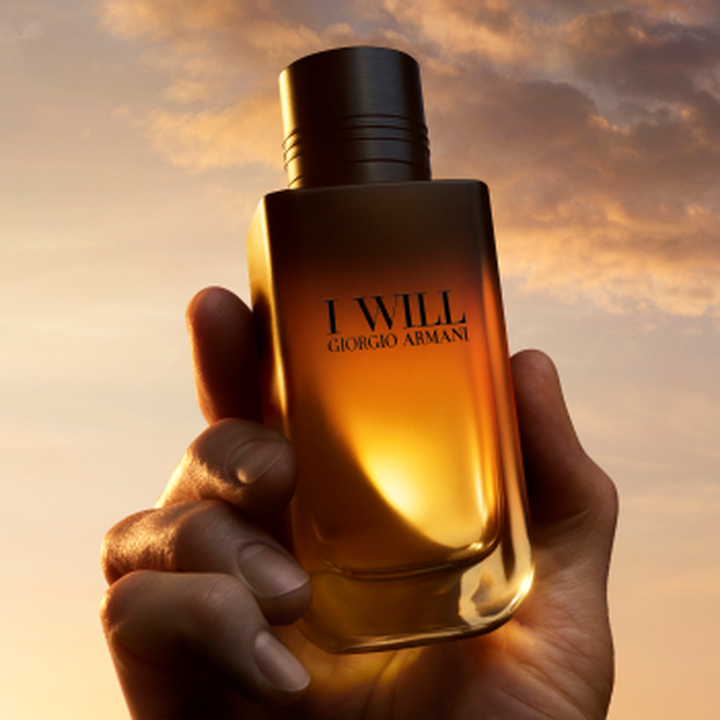 Eine Hand halt den Duftflakon I WILL von Giorgio Armani vor einem goldenen Sonnenuntergangshimmel.