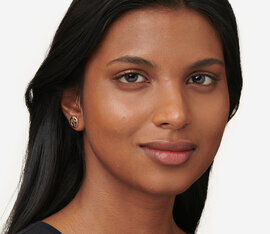 A close-up portrait of a woman with long black hair, smooth skin, and subtle makeup, smiling softly while looking directly at the camera. She is wearing small earrings.