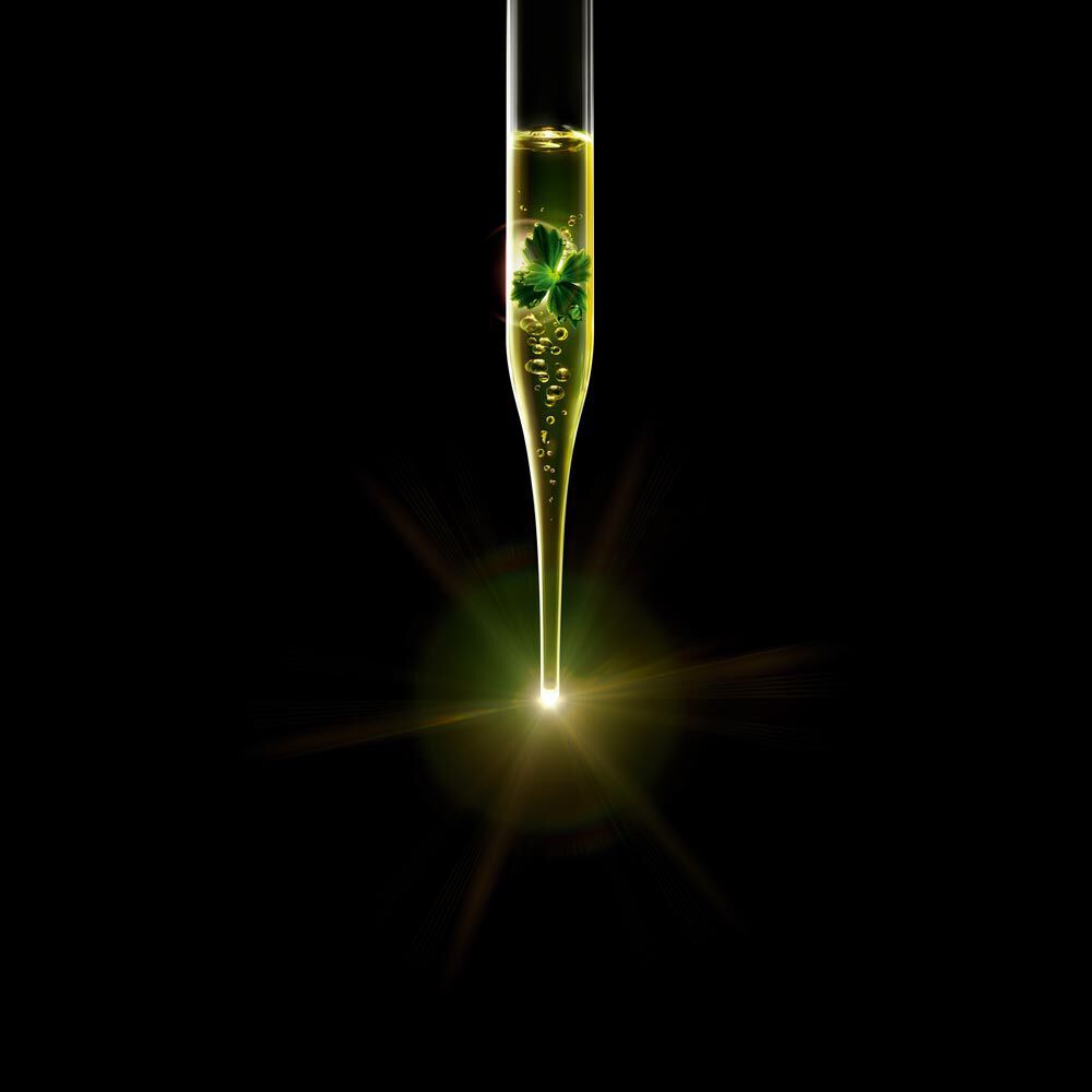 A dropper filled with a golden liquid containing green leaves, suspended above a soft glowing light against a black background.