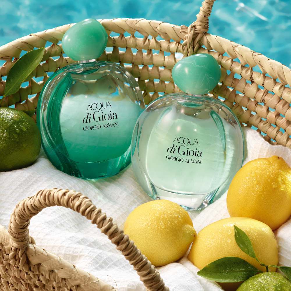 Two bottles of Acqua di Gioia perfume by Giorgio Armani are nestled in a woven basket with a white cloth, accompanied by a lemon and a lime, set against a backdrop of blue water.