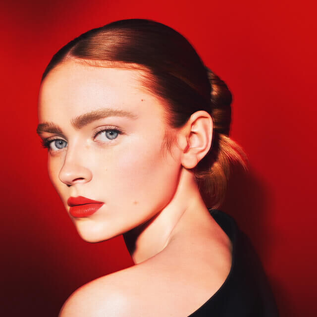 Eine Frau mit blonden Haaren und blauen Augen steht vor einem roten Hintergrund. Sie hat einen eleganten Haarschnitt und tragt rote Lippenfarbe.