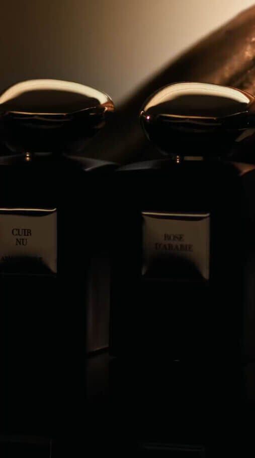 Mille et une nuits collection video showcasing three black Armani/Prive fragrance bottles, labeled "OUD ROYAL", "CUIR NU", and "ROSE D'ARABIE", with gold caps and accents, arranged on a reflective surface alongside elements like vanilla pods, a piece of golden-dusted wood, and a golden-dusted fig.