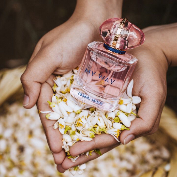 A pair of hands holding a bottle of Giorgio Armani's "My Way" perfume surrounded by fragrant white flowers.