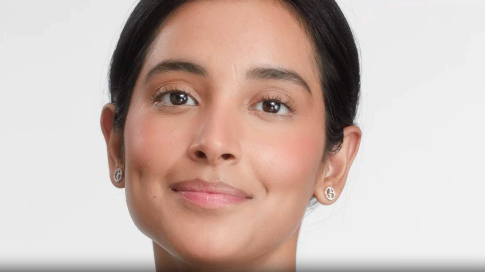 A close-up portrait of a woman with dark hair pulled back, smiling gently, and wearing small earrings. She has a neutral background and a natural makeup look.