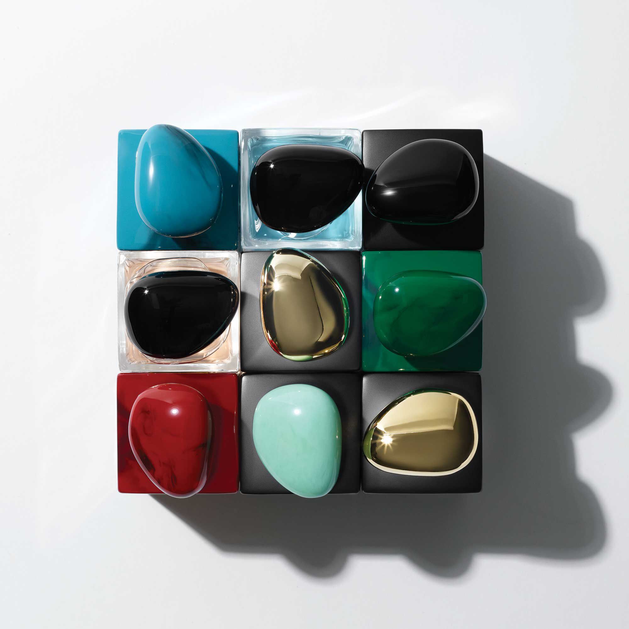 A symmetrical arrangement of nine colorful, smooth, pebble-shaped objects sitting atop black cubes, featuring shades of turquoise, black, green, red, and gold against a light background.