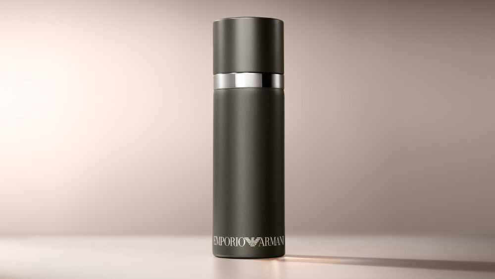A matte black cylindrical Emporio Armani "He" fragrance bottle is presented on a warm, light brown background in a horizontal aspect ratio.