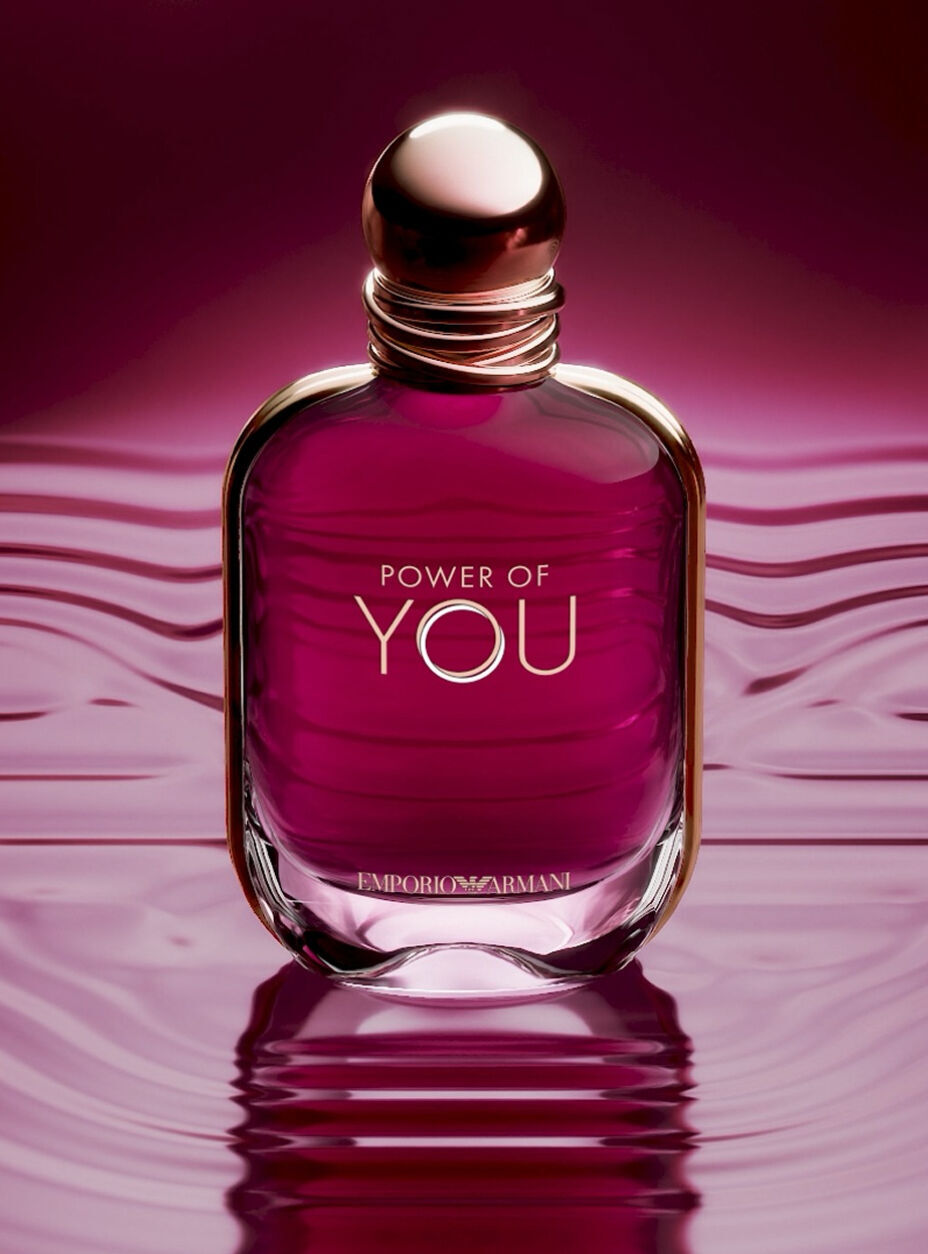 Eine Nahaufnahme des Parfumflakons „Power of You“ von Emporio Armani. Der rubinfarbene Glasflakon steht zentral auf einer reflektierenden, dunkelmagentafarbenen Flussigkeitsoberflache, die sanfte Wellen zeigt. Der Flakon hat einen abgerundeten goldenen Verschluss und die in Gold aufgedruckten Worte „POWER OF YOU“ und „EMPORIO ARMANI“. Der Hintergrund ist ein weicher, verschwommener Farbverlauf aus tiefem Rosa und Lila. Am unteren Rand steht in weißem Text: „Einfuhrung eines neuen ikonischen Flakons: ein rubinfarbenes Juwel“, gefolgt von einem „ENTDECKEN“-Link und einem Pause-Symbol.