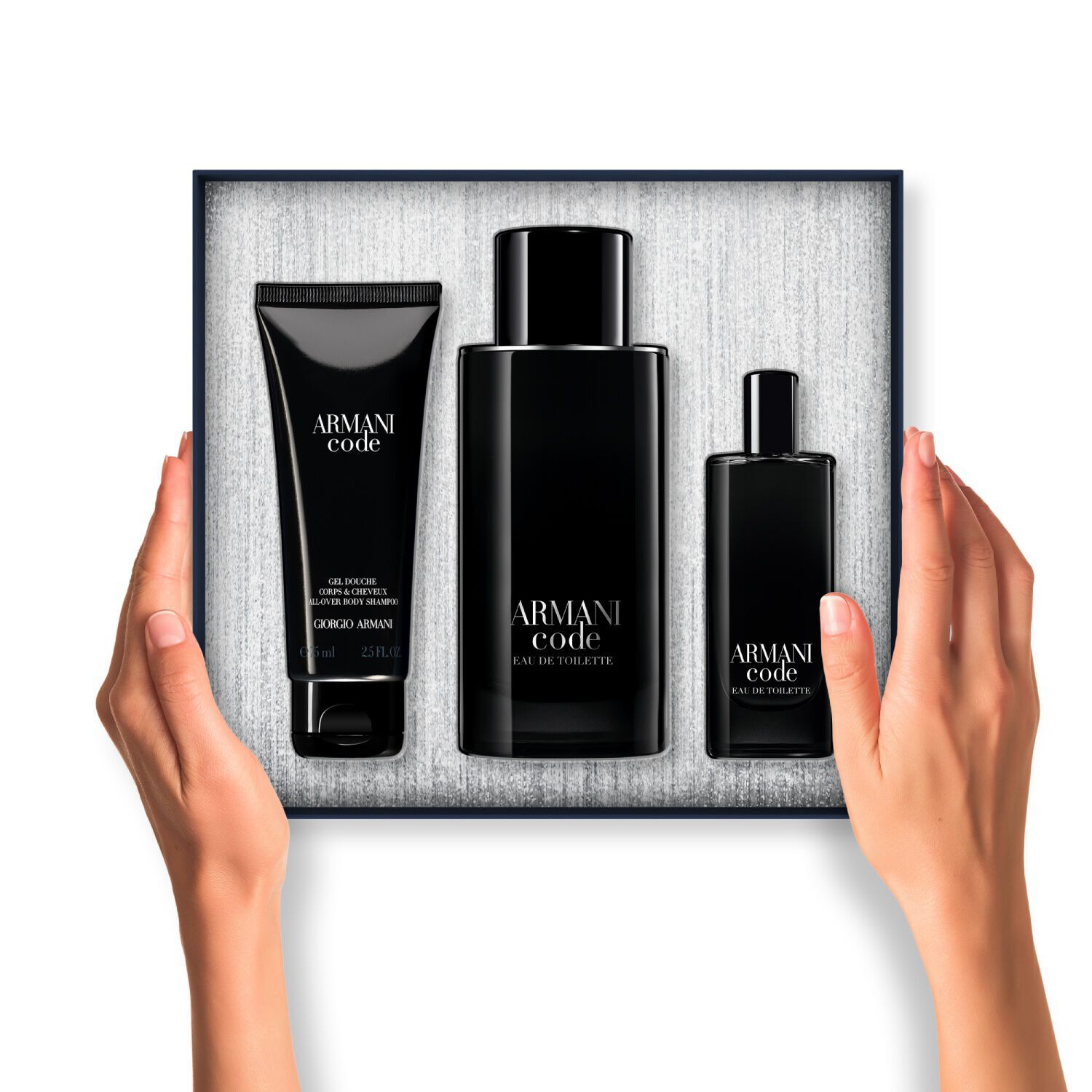 Ein Paar Hande halt eine offene Armani Code Geschenkbox. Darin sind drei schwarz verpackte Produkte (Korpergel, großes Eau de Toilette und Reisegroße Eau de Toilette) ordentlich auf einem texturierten silbernen Hintergrund arrangiert.