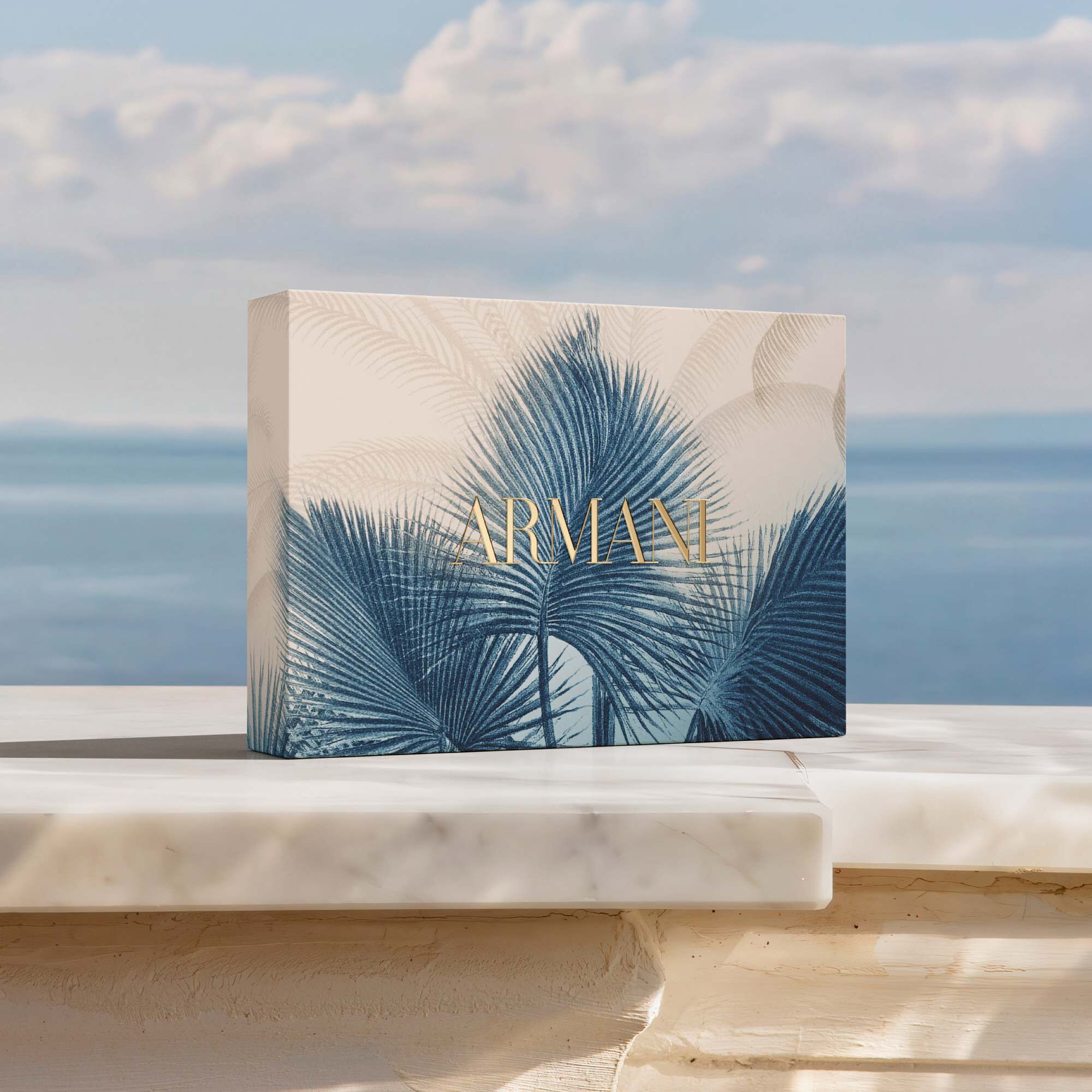 Giorgio Armani Acqua di Gio summer gift box featuring an elegant blue palm leaf design and gold logo. A luxury gift set presented on a sunlit marble terrace overlooking the Mediterranean sea.