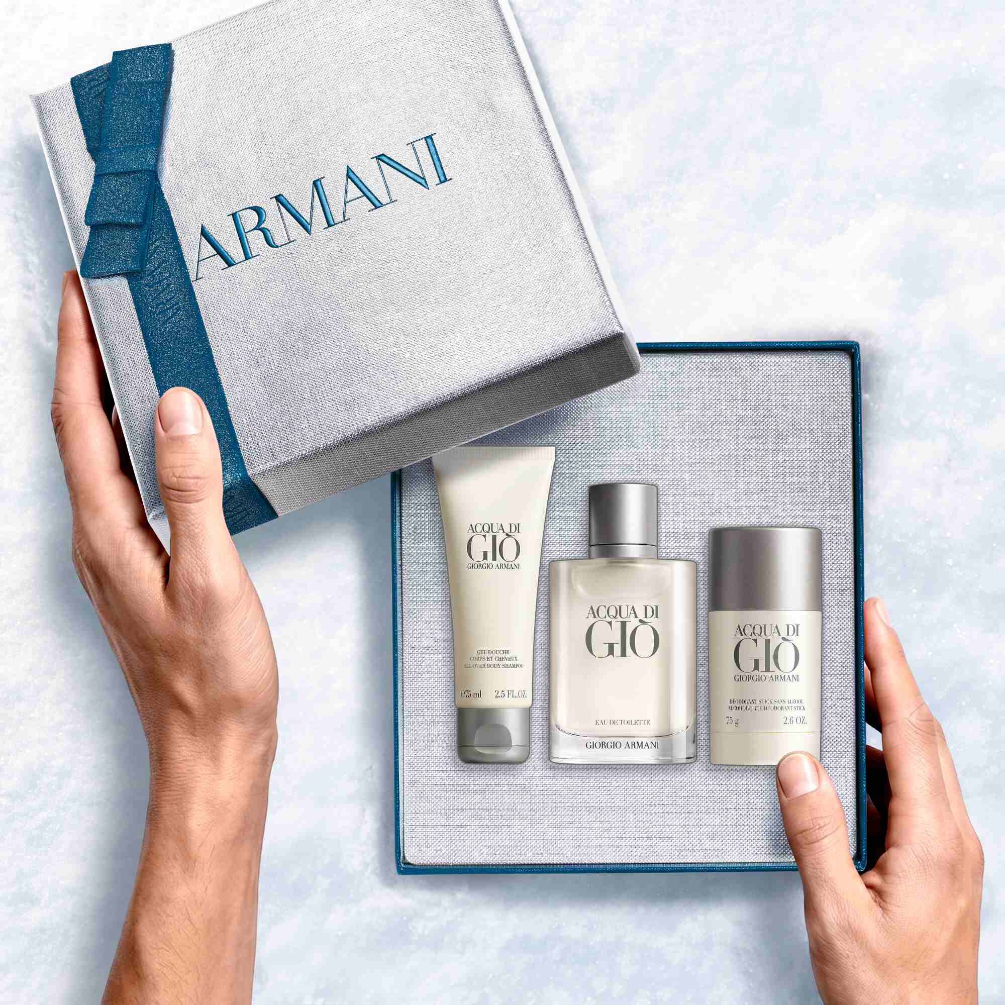 Giorgio Armani Acqua di Gio Eau de Toilette 100ml gift set — hands unveiling the signature bottle, All-Over Body Shampoo 75ml and Deodorant Stick 75g from within the open Armani gift box, an intimate unboxing moment.