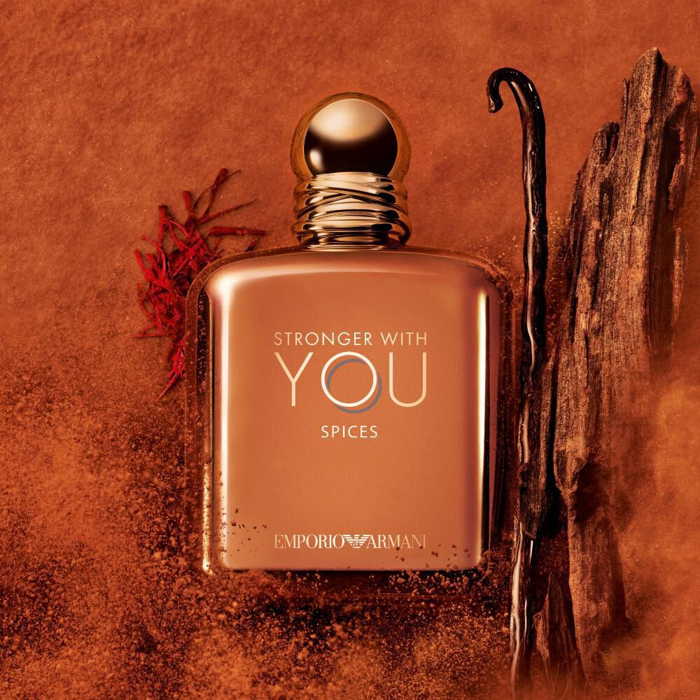 Flakon von ARMANI STRONGER WITH YOU SPICES mit goldenem Verschluss, platziert zwischen Vanilleschoten und Safran auf einem orangefarbenen, strukturierten Hintergrund. Das Bild unterstreicht die wurzigen Gourmand-Noten des Duftes.