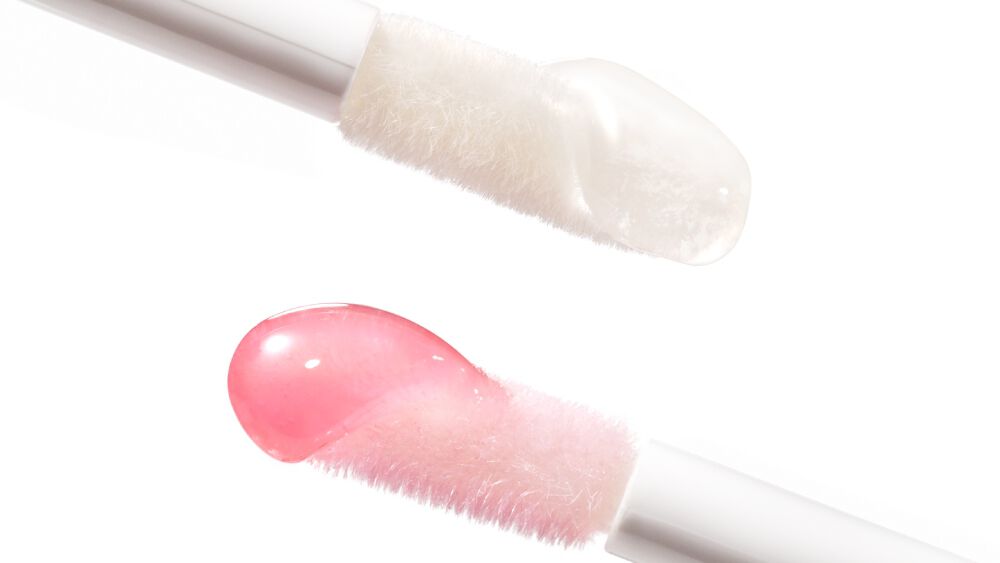 Close-up of two lip gloss applicators, one with a clear gloss and the other with a pink gloss, against a white background.
